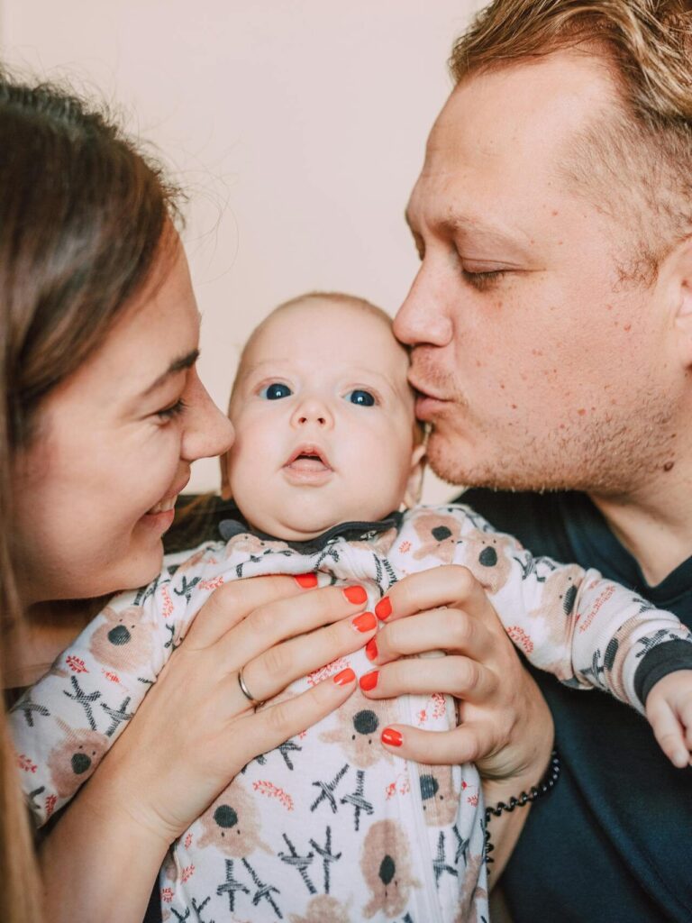 Loving parents embrace and kiss their newborn baby indoors.